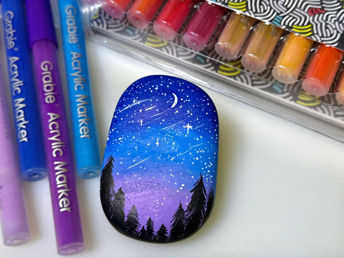 Hand-painted rock with night forest sky, acrylic markers and colored paint pens nearby.