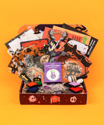 Halloween-Themed Grabie Scrapbook Club Box with assorted spooky papers, stamps, ink pads, and crafting supplies on orange background