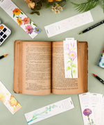 Open book with a floral bookmark surrounded by watercolor art supplies for The Watercolor Beginner‘s Bundle