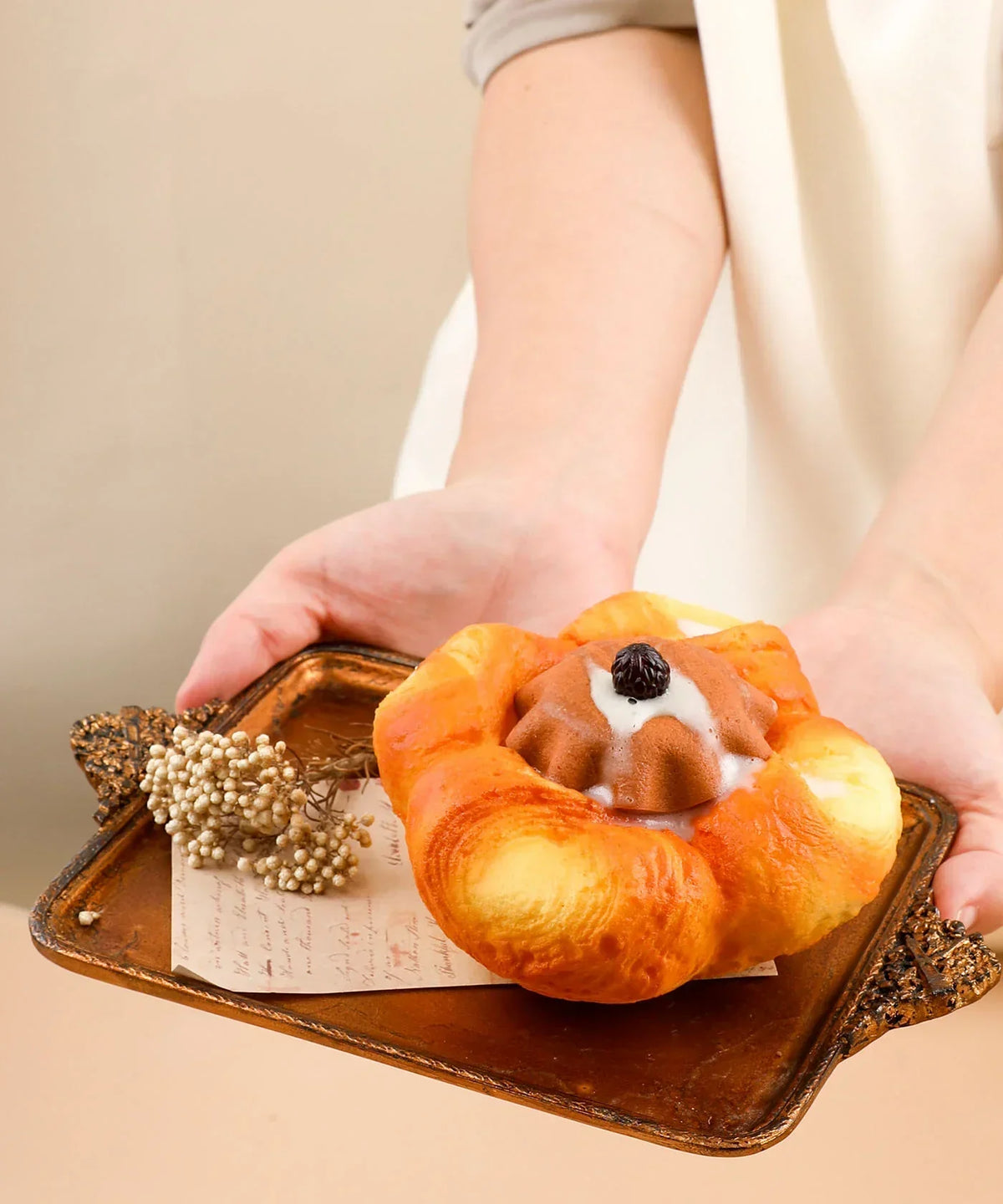 Hands holding a tray with Feel-Good Stress Relief Foam Squishy - Chocolate Flower Bread resting on decorative paper and dried flowers.