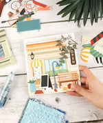 Hand holding cozy decorated scrapbook page with art supplies, featuring Cozy Pop Stores-Themed Grabie Scrapbook Club Box