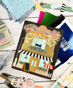Art supplies and colorful scrapbook elements arranged around a Cozy Pop Stores-Themed Grabie Scrapbook Club Box cover.