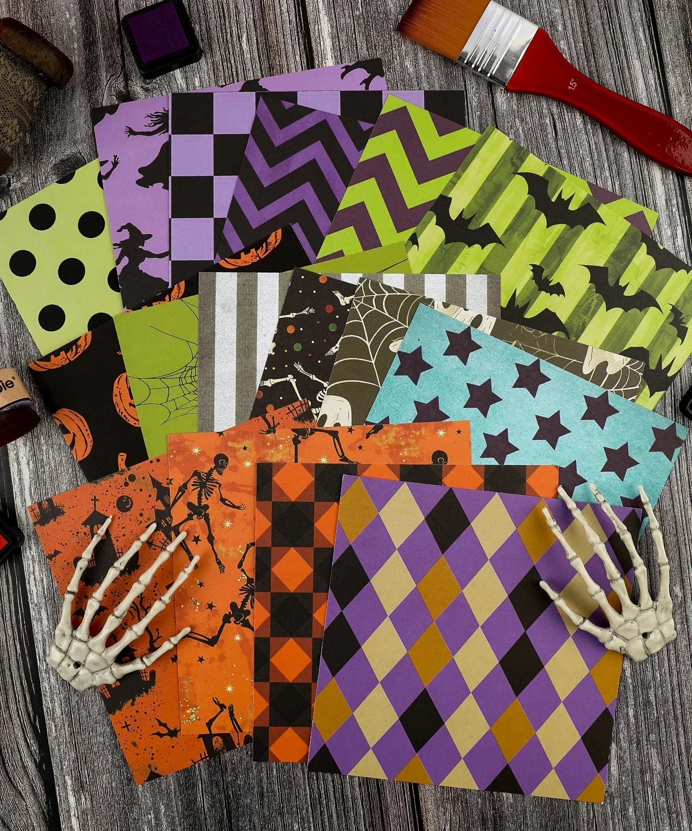 Colorful Halloween-themed patterned papers arranged on wooden surface with paintbrush and skeleton hands, 16 Sheets Grabie Exclusive Halloween Party Paper Set