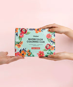 Hands exchanging the Floral-Themed Watercolor Coloring Club Box with bright floral artwork on a pastel background