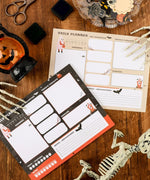 30 sheets Halloween Exclusive Daily Planner with spooky illustrations on wooden desk surrounded by craft supplies and skeleton props