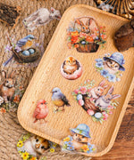 60 Pcs Grabie Exclusive The Magic Of Easter Sticker Set with colorful bunnies, chicks, birds, and eggs on a wooden tray