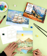 Hands painting a landscape watercolor scene with art supplies and the Landscape-Themed Watercolor Coloring Club Box nearby