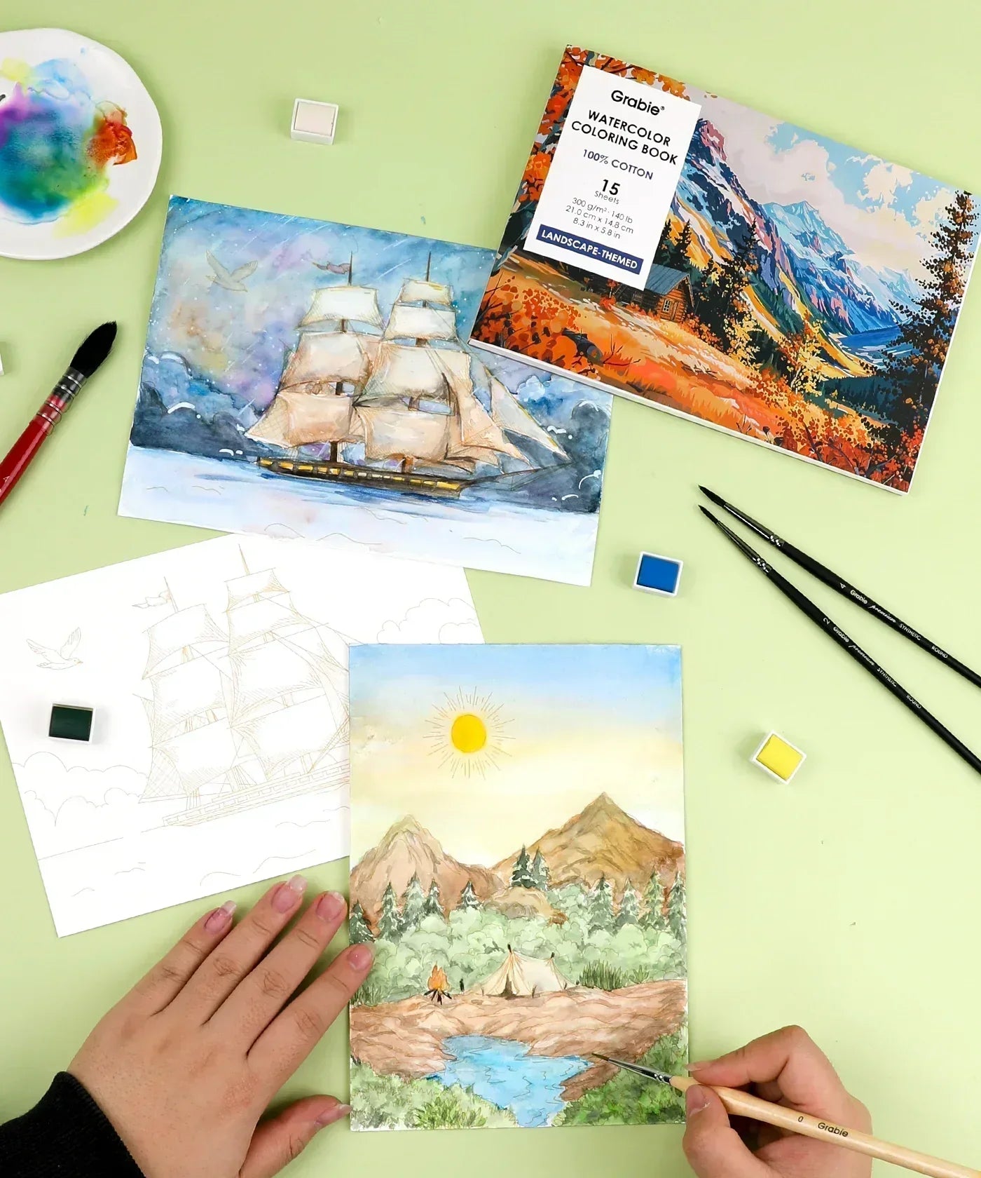 Hands painting a landscape watercolor scene with art supplies and the Landscape-Themed Watercolor Coloring Club Box nearby