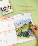 Affirm Your World 4-Week Workbook open with watercolor landscape painting and brushes on a green surface.