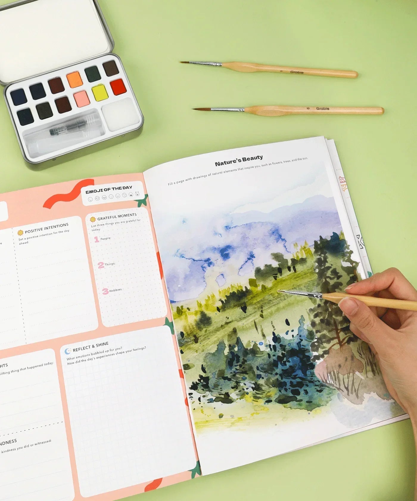 Affirm Your World 4-Week Workbook open with watercolor landscape painting and brushes on a green surface.