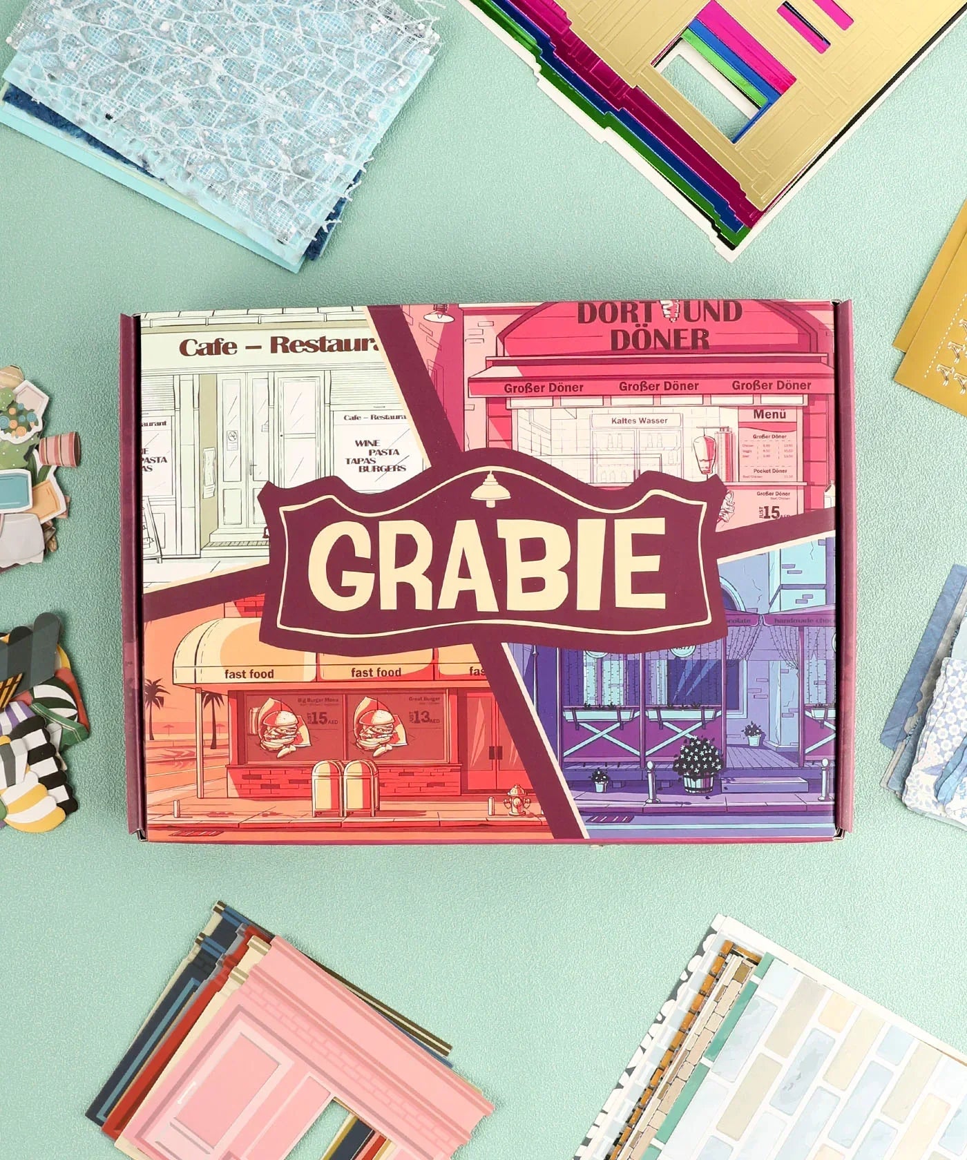 Cozy Pop Stores-Themed Grabie Scrapbook Club Box surrounded by colorful paper scrapbooking supplies and cutouts.