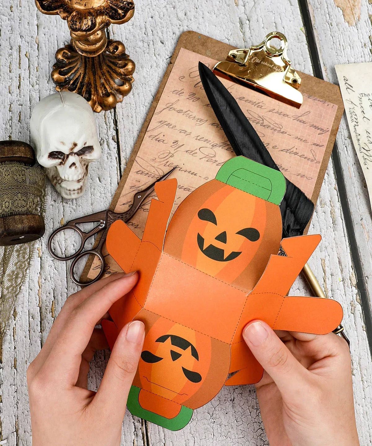 Hands assembling paper pumpkin craft with scissors and skull, part of the 3 Sheets Grabie Exclusive Halloween DIY Kit