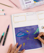 Person coloring a starry sky page inside the Be You And Love You 28-day Workbook with markers and pens nearby