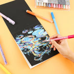 Hand using Extra Fine Tip Acrylic Paint Marker Set of 48 to create colorful art on black paper with markers nearby