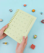 Hands holding a pale yellow 2-IN-1 POPIN Notebook with a calendar design on the cover against a blue background