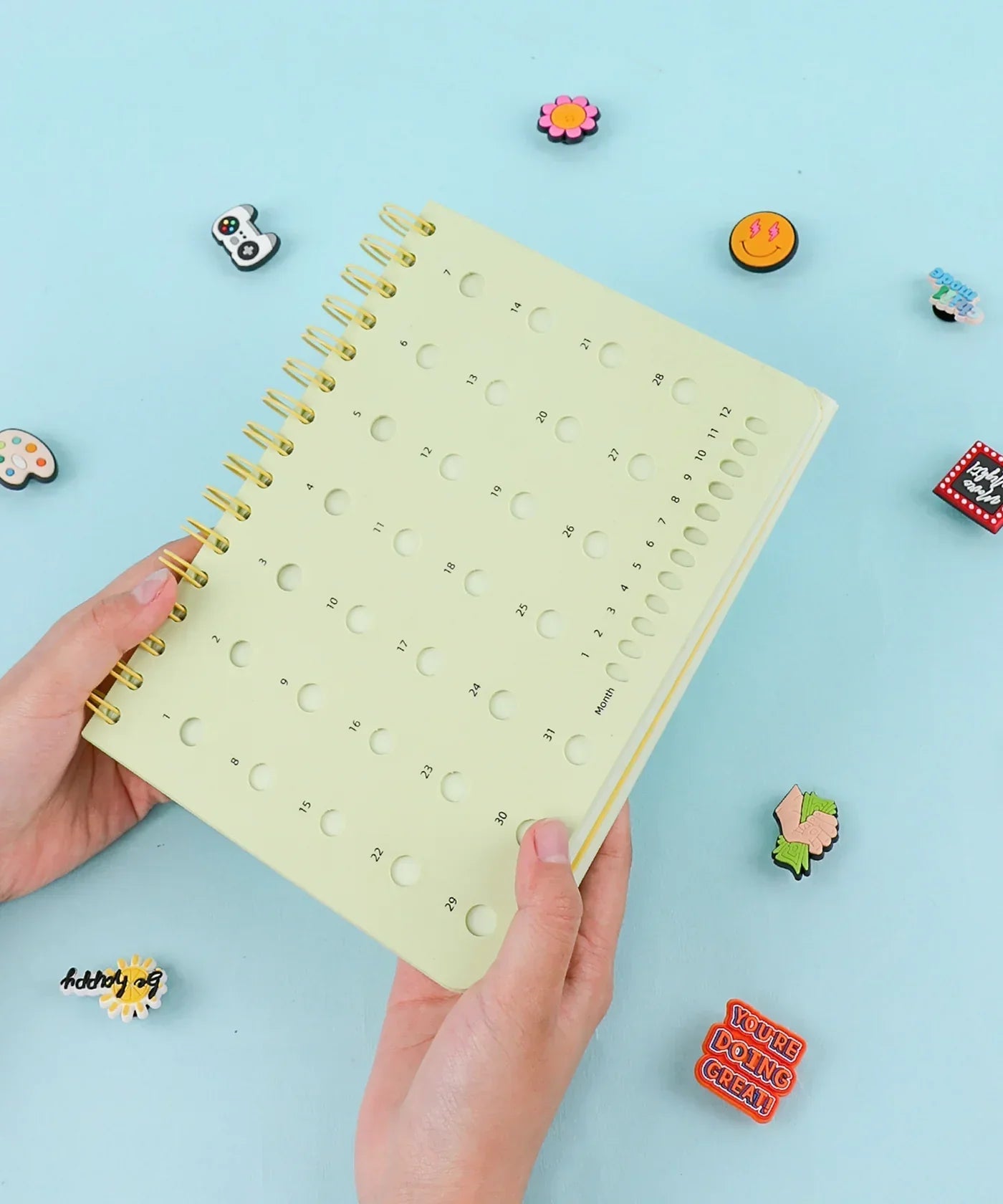 Hands holding a pale yellow 2-IN-1 POPIN Notebook with a calendar design on the cover against a blue background