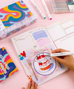 Person coloring a strawberry cake illustration in the Be You And Love You 28-day Workbook with markers and pens nearby