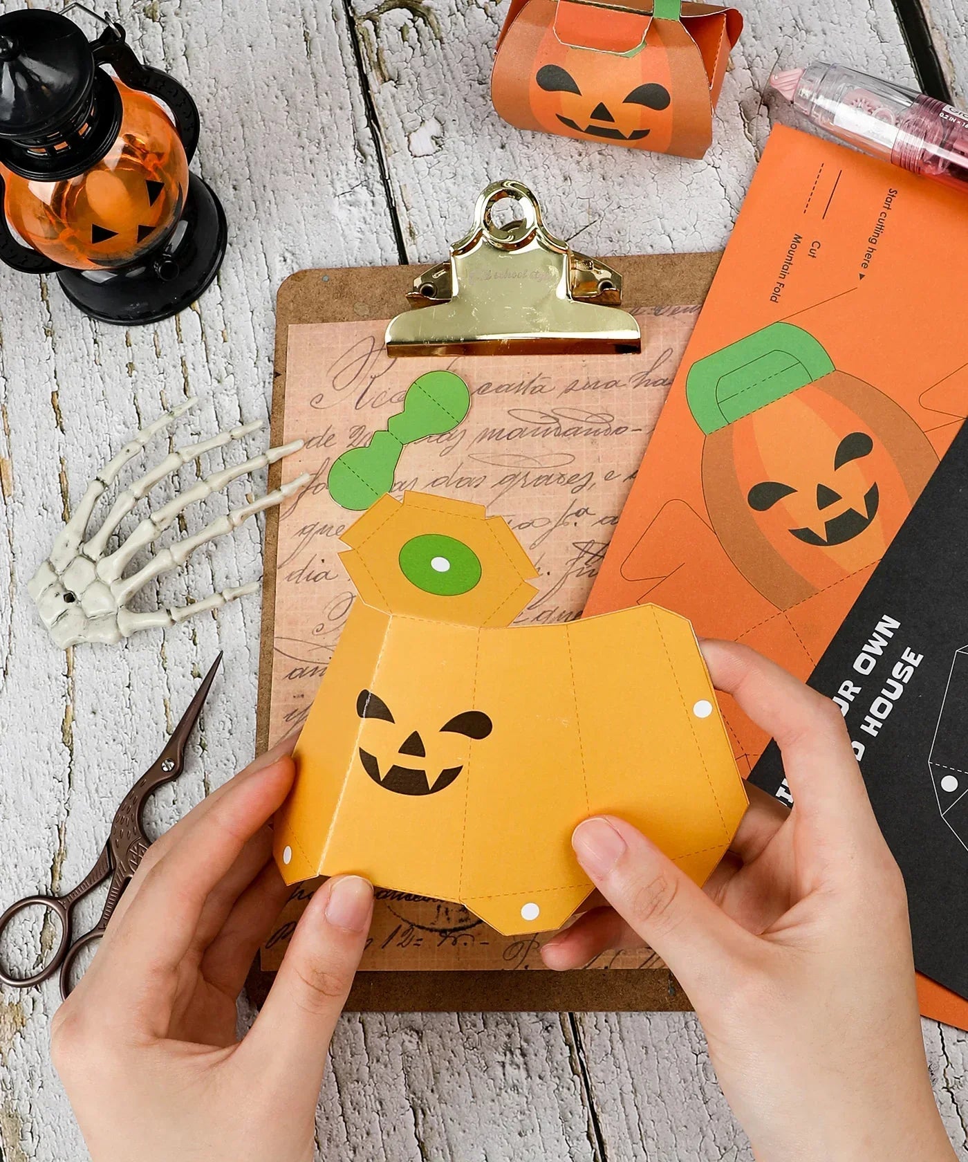 Hands assembling a paper pumpkin craft from the 3 Sheets Grabie Exclusive Halloween DIY Kit on a wooden table.
