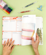 Hands flipping through Affirm Your World 4-Week Workbook with colorful markers, pens, and clipped binder on green surface