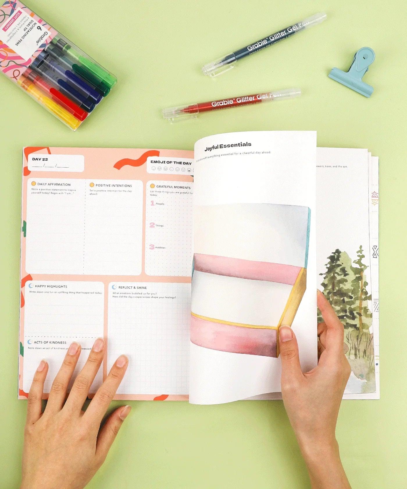 Hands flipping through Affirm Your World 4-Week Workbook with colorful markers, pens, and clipped binder on green surface
