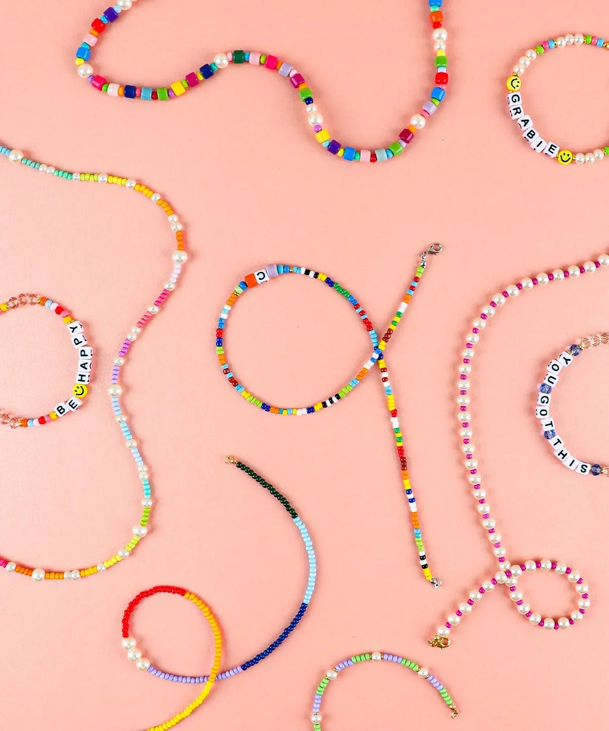 Colorful handmade beaded jewelry including bracelets and necklaces with letters on a pink background featuring Grabie Bead Box