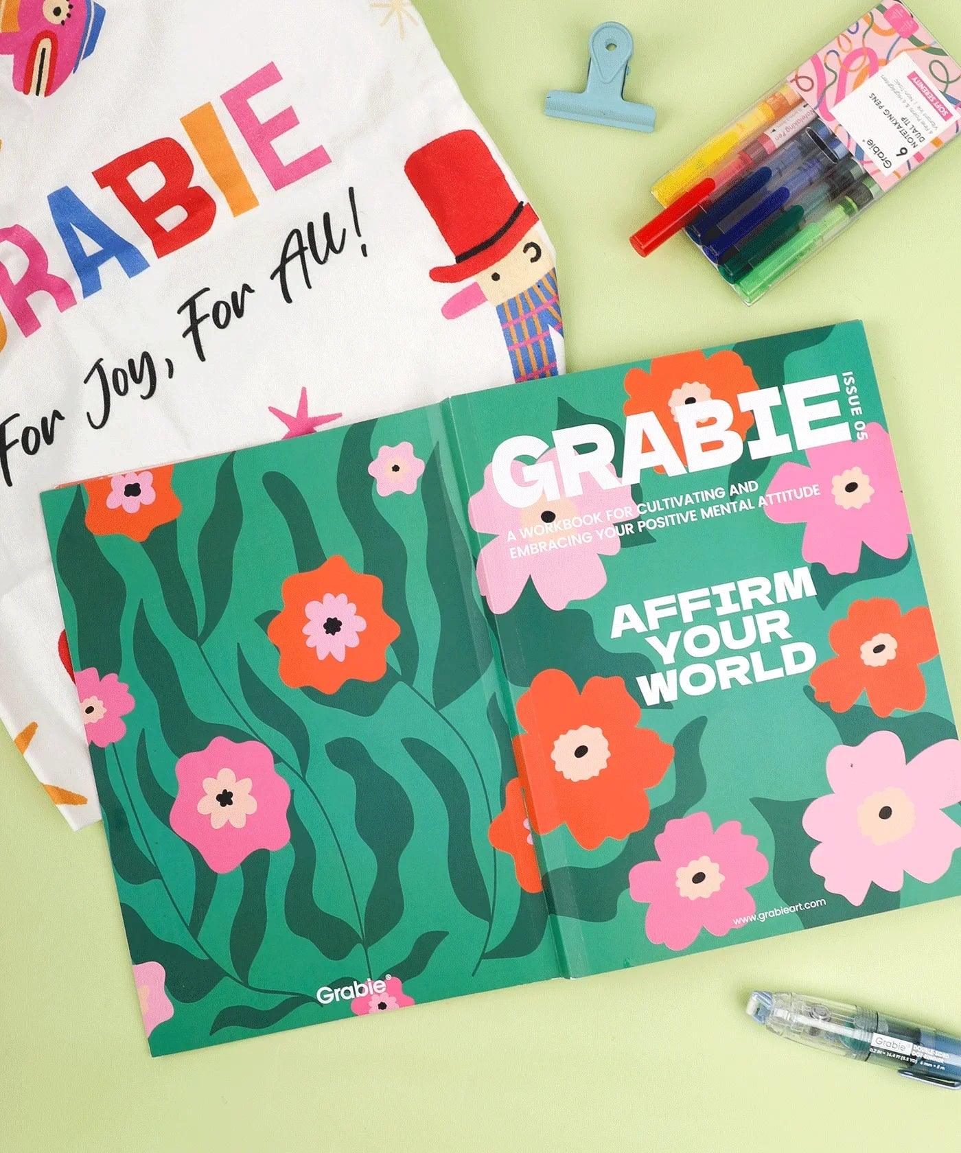 Open Affirm Your World 4-Week Workbook with floral design next to colorful pens and a branded tote bag on green surface