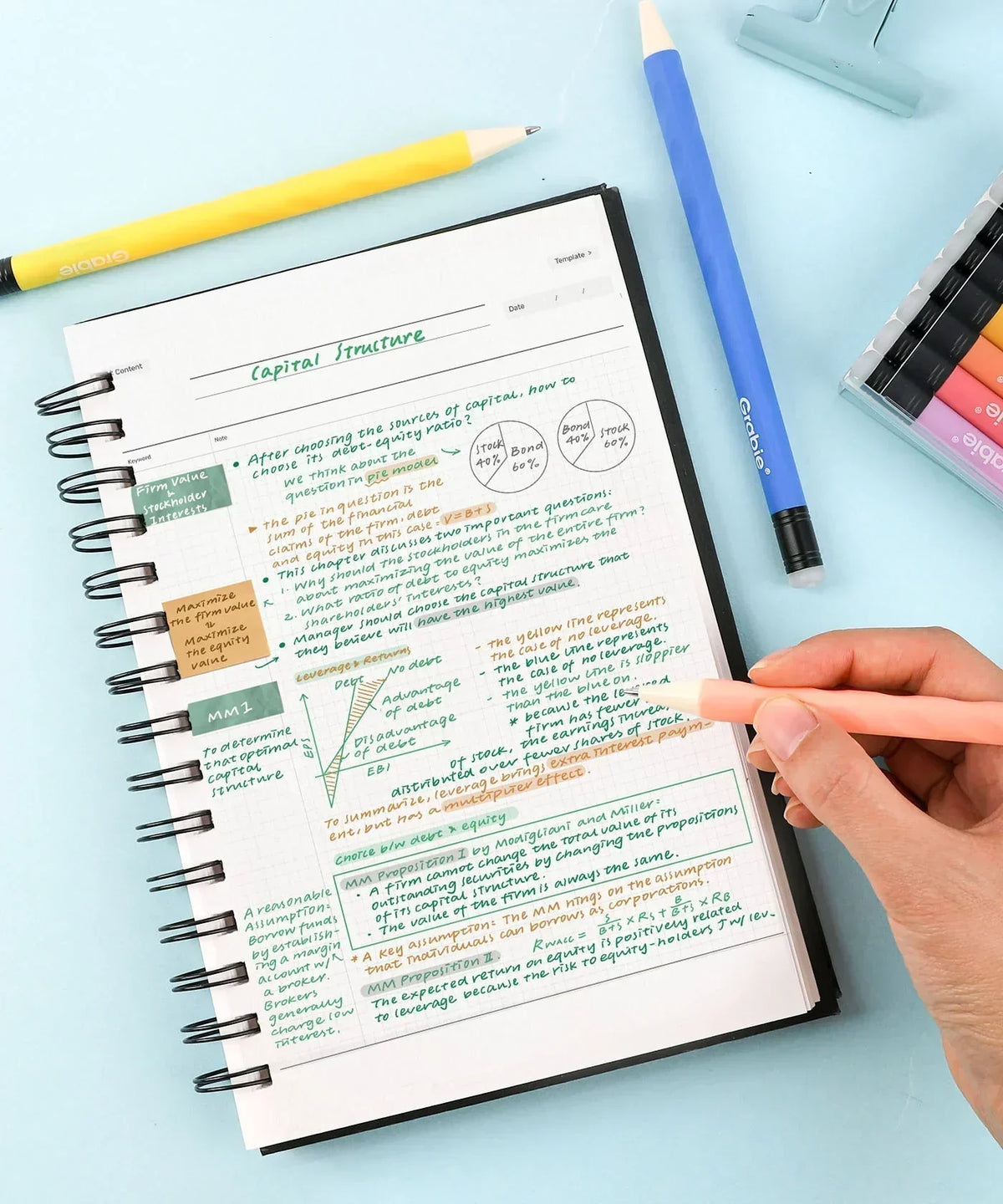 Hand holding a pen writing colorful notes in a spiral notebook alongside the Medium Point Erasable Gel Pen Set and pencils.