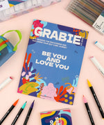 Be You And Love You 28-day Workbook surrounded by colorful art supplies including markers and pens on a pastel surface