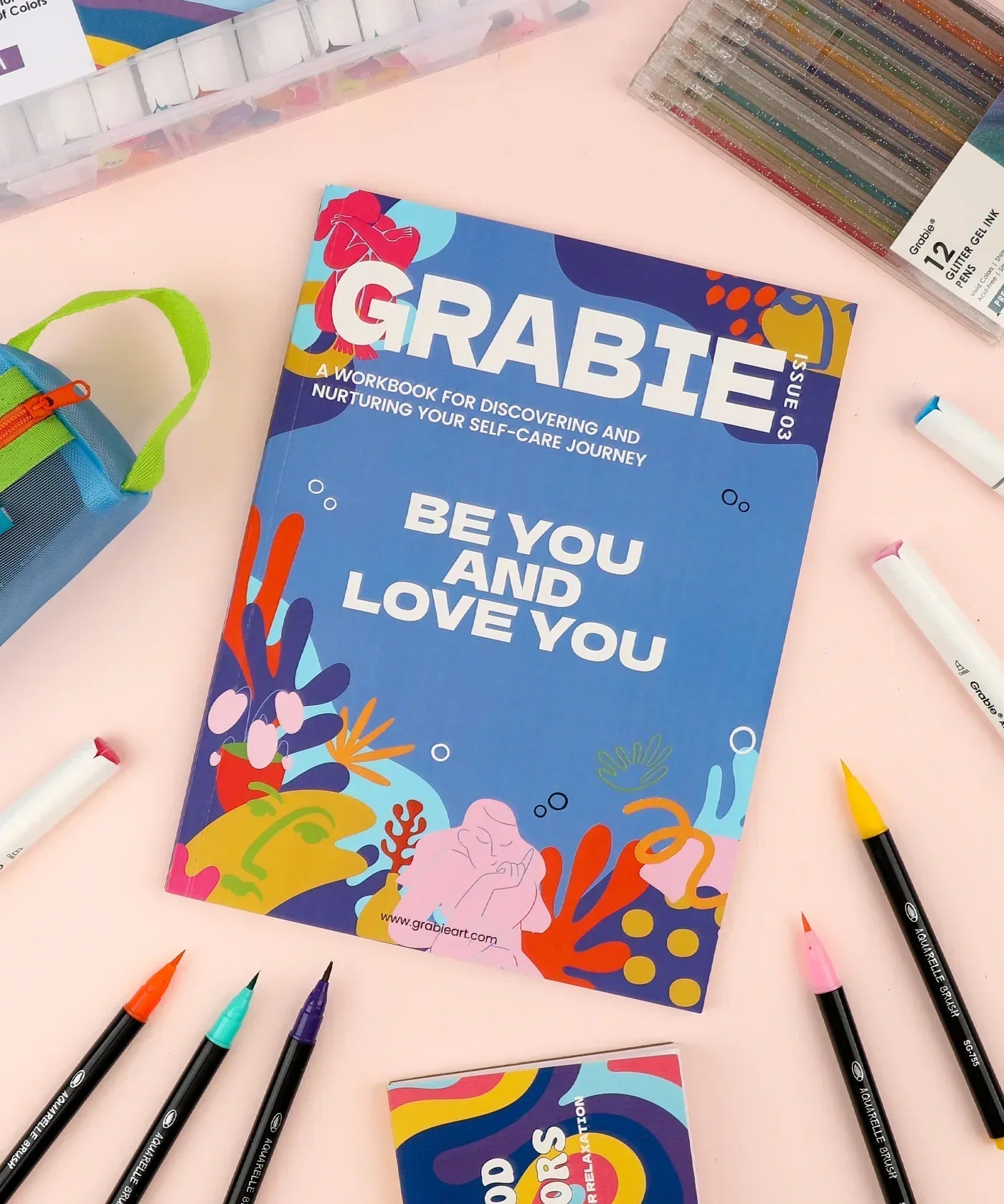 Be You And Love You 28-day Workbook surrounded by colorful art supplies including markers and pens on a pastel surface