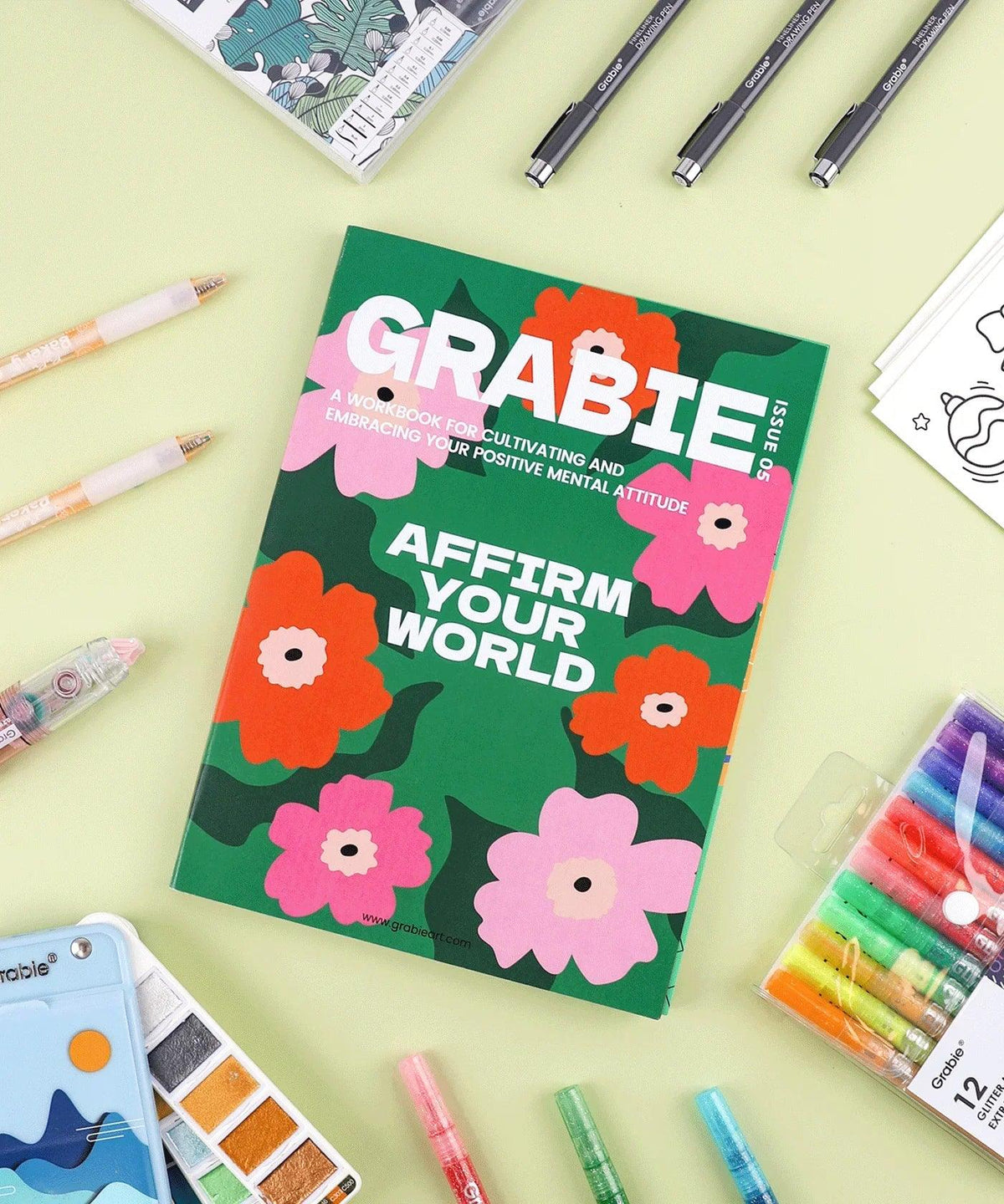 Affirm Your World 4-Week Workbook with colorful floral cover surrounded by art supplies on a light green background