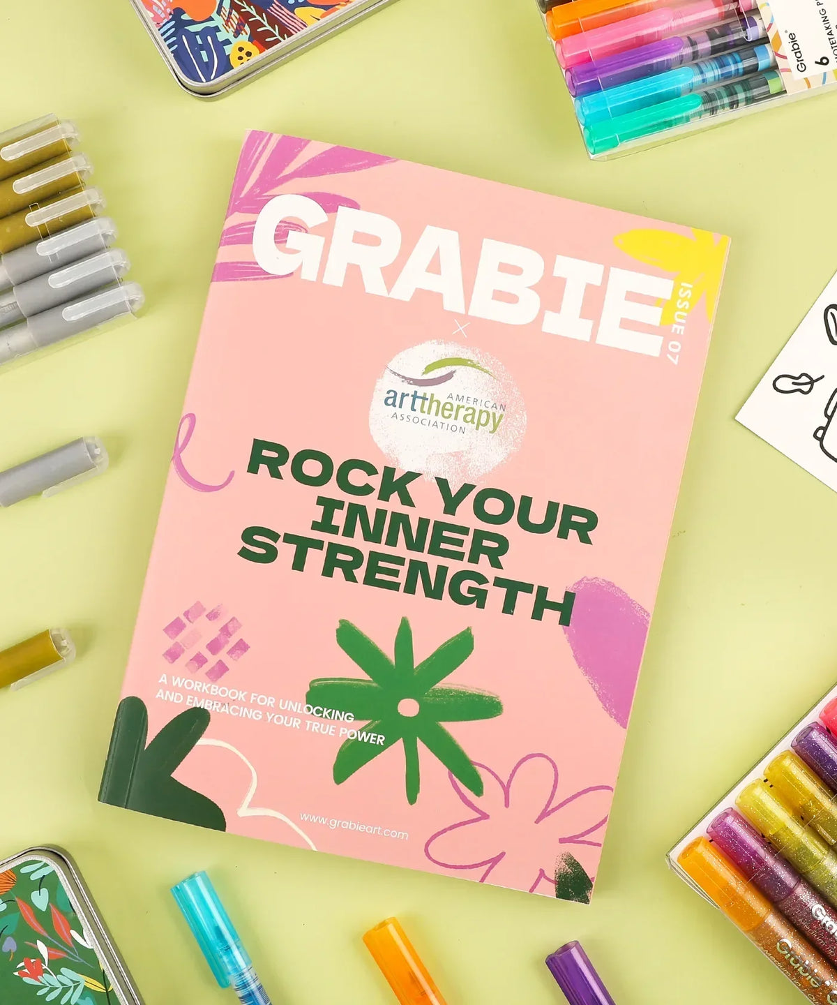 Grabie x AATA Rock Your Inner Strength 4-Week Workbook surrounded by colorful markers and art supplies on a yellow background