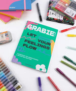Let Your Feelings Flow Grabie Art Club Box surrounded by colorful markers, pens, and art supplies on white surface