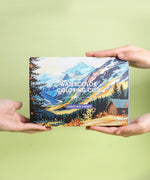 Hands holding a Landscape-Themed Watercolor Coloring Club Box featuring mountain and cabin art supplies set