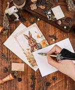 Grabie Exclusive 6 Pack Postcards With 6 Matching Envelopes featuring a vintage rabbit illustration on wooden table with writing hand.