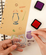 Hands using Happy Halloween Clear Photopolymer Stamps to create colorful stamped images on a brown notebook page next to ink pads