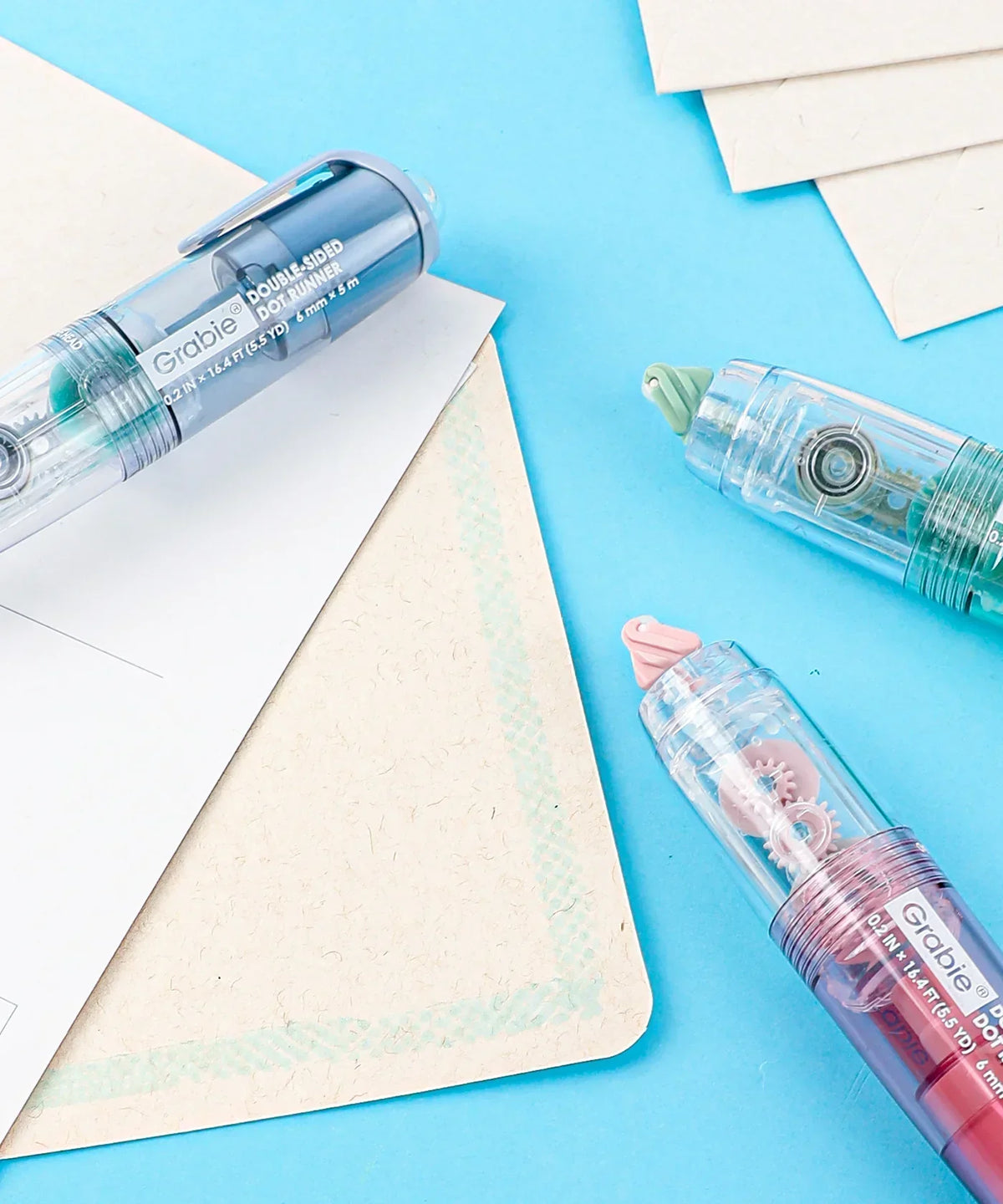 Three Double-Sided Adhesive Dot Runner rollers with transparent casing on blue surface and paper sheets nearby