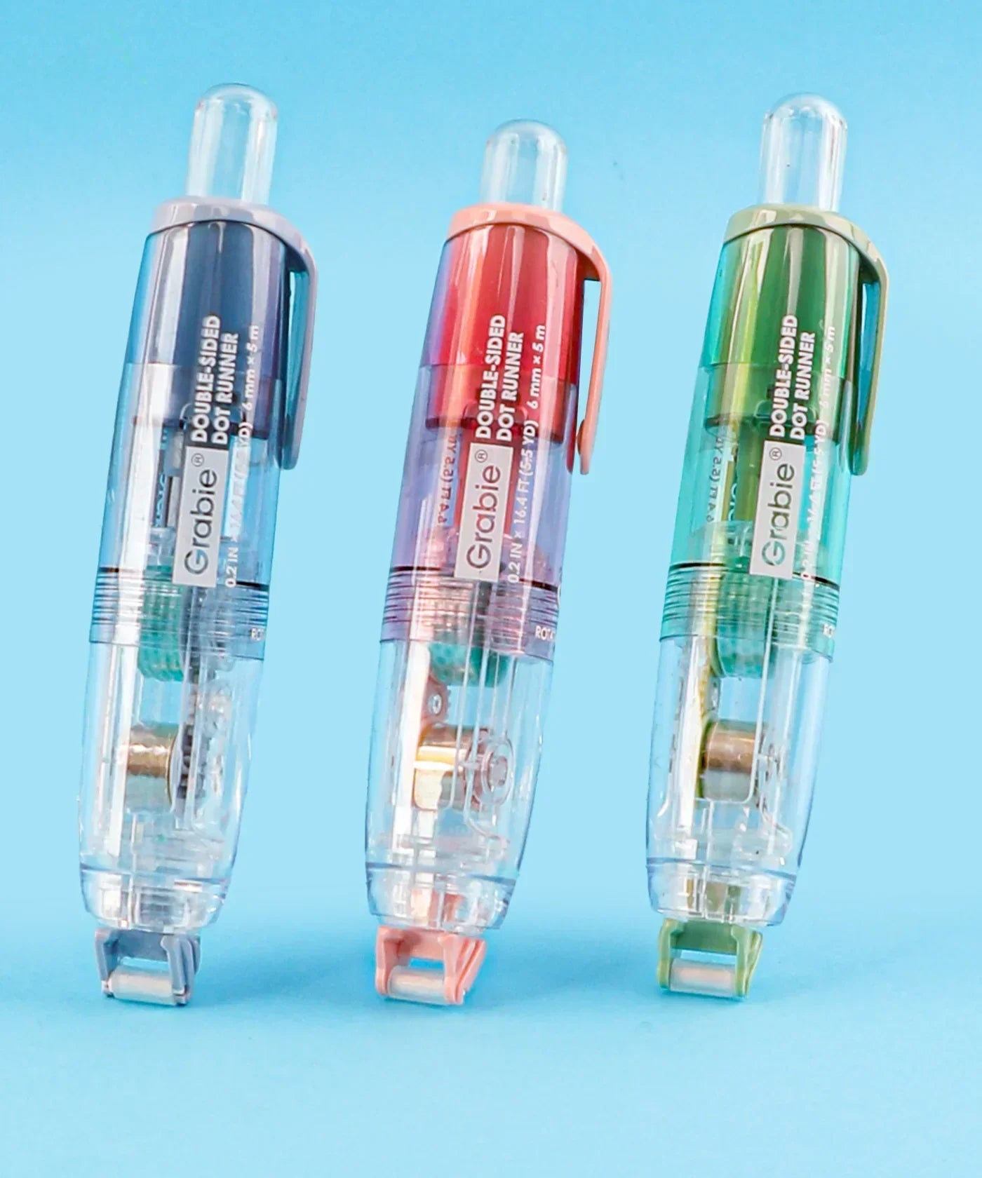 Three Grabie Double-Sided Adhesive Dot Runner dispensers in blue, pink, and green on a blue background