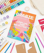 Unplug And Go Outside 30-day Workbook surrounded by colored pencils, watercolor paints, brushes, and craft supplies on white background.