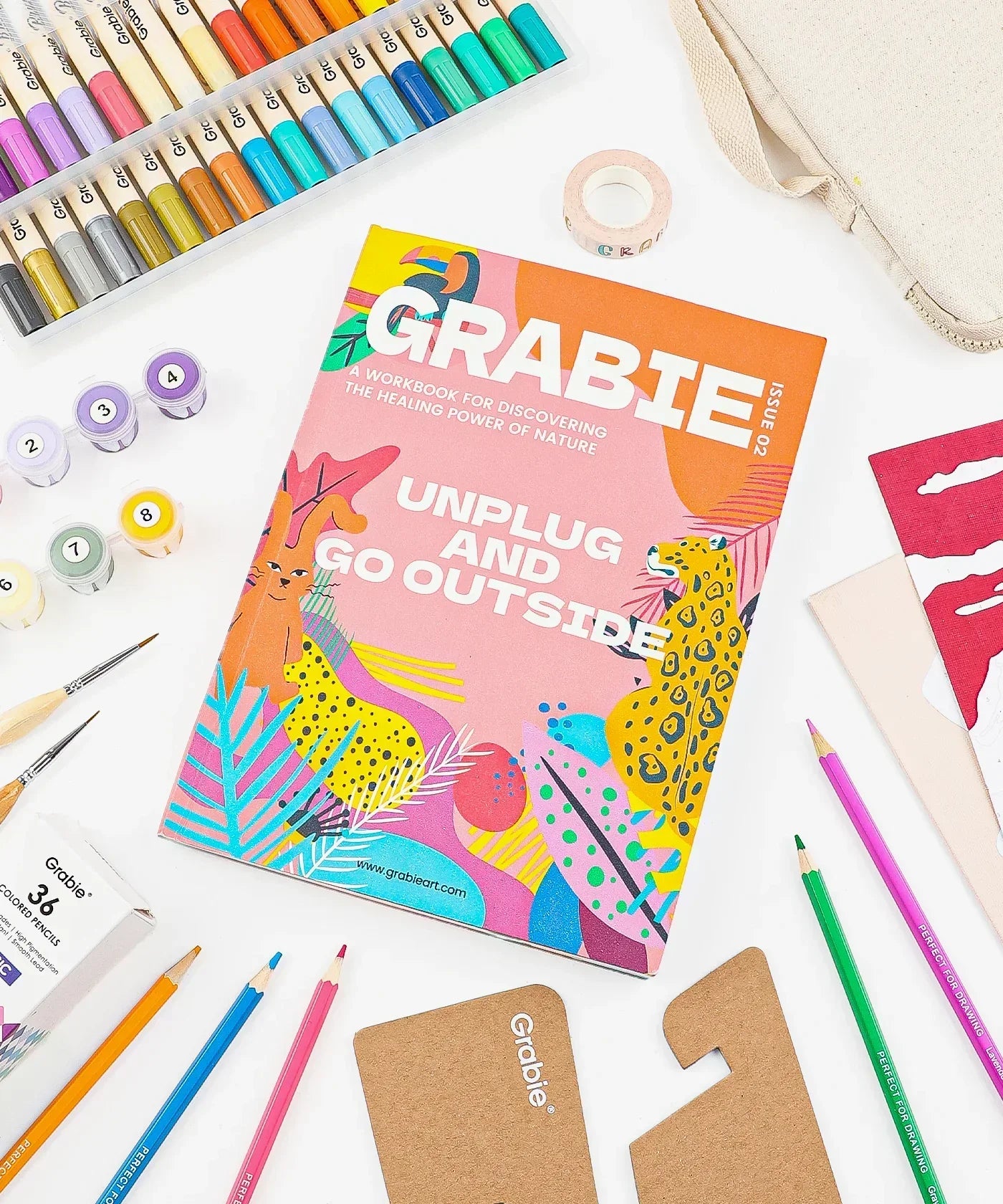 Unplug And Go Outside 30-day Workbook surrounded by colored pencils, watercolor paints, brushes, and craft supplies on white background.