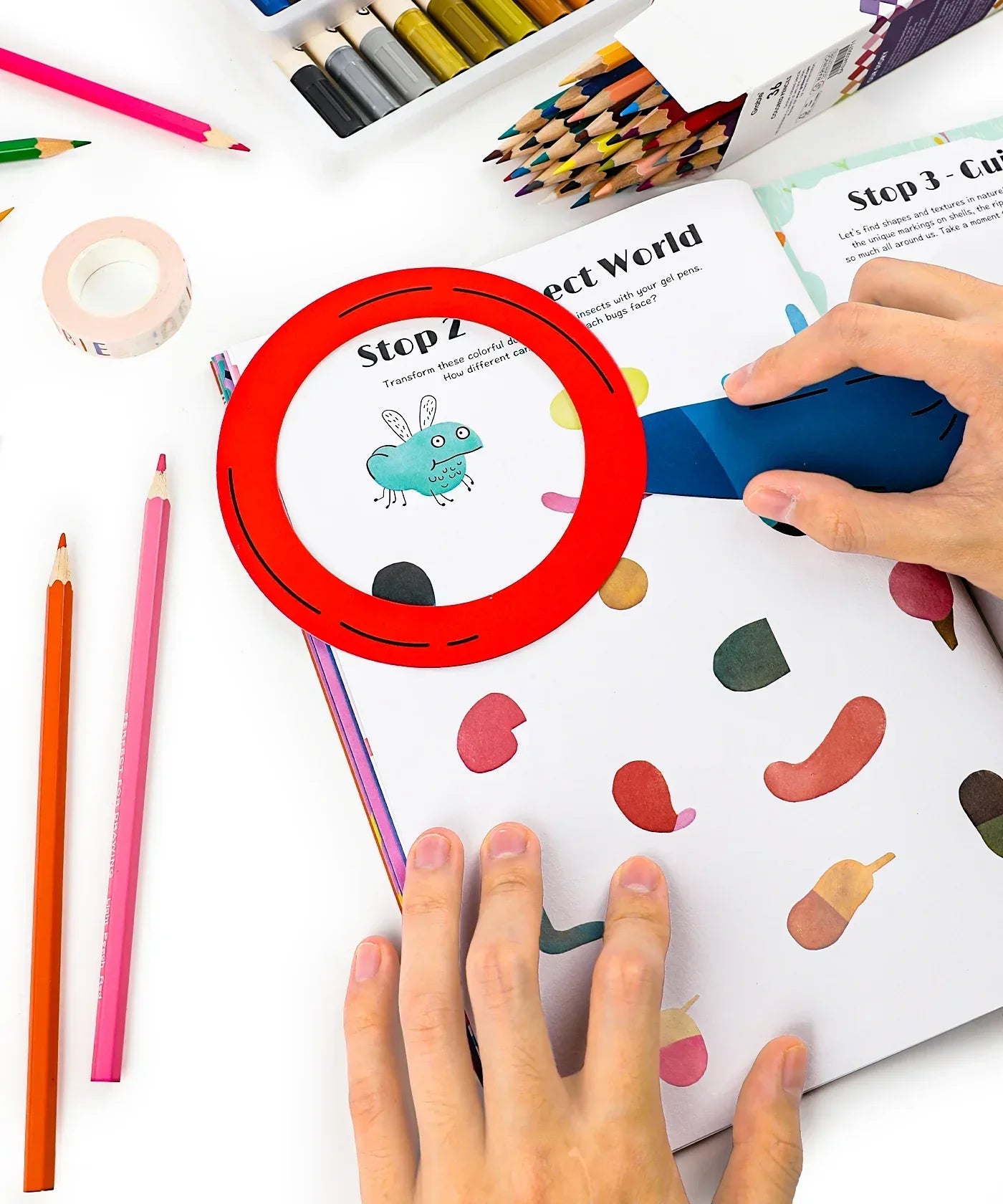 Hands using a magnifying glass on colorful pages of Unplug And Go Outside 30-day Workbook with art supplies nearby