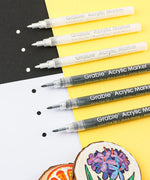 Grabie Acrylic Paint Marker Set with black and white markers showing paint dots on black, white, and yellow paper with wood slice art.