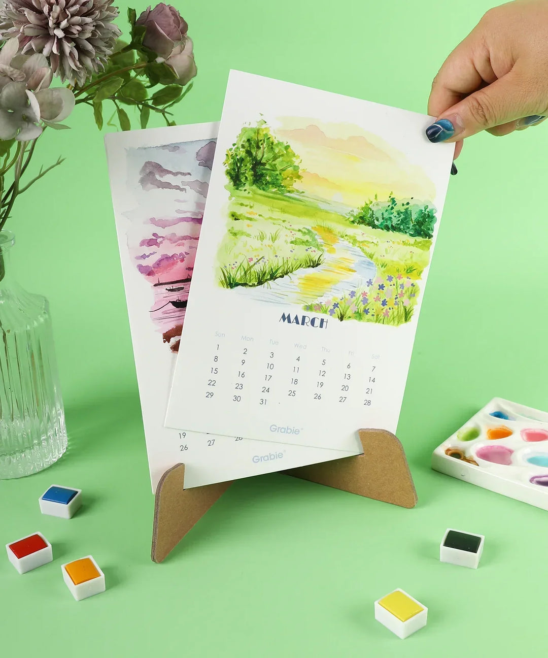 Hand holding March page of 12-Month Watercolor Calendar Set For 2026 with floral watercolor art on green background