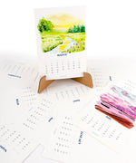 2026 Watercolor Calendar & Creative Tools Set with scenic watercolor month pages displayed on a stand and spread out on a surface
