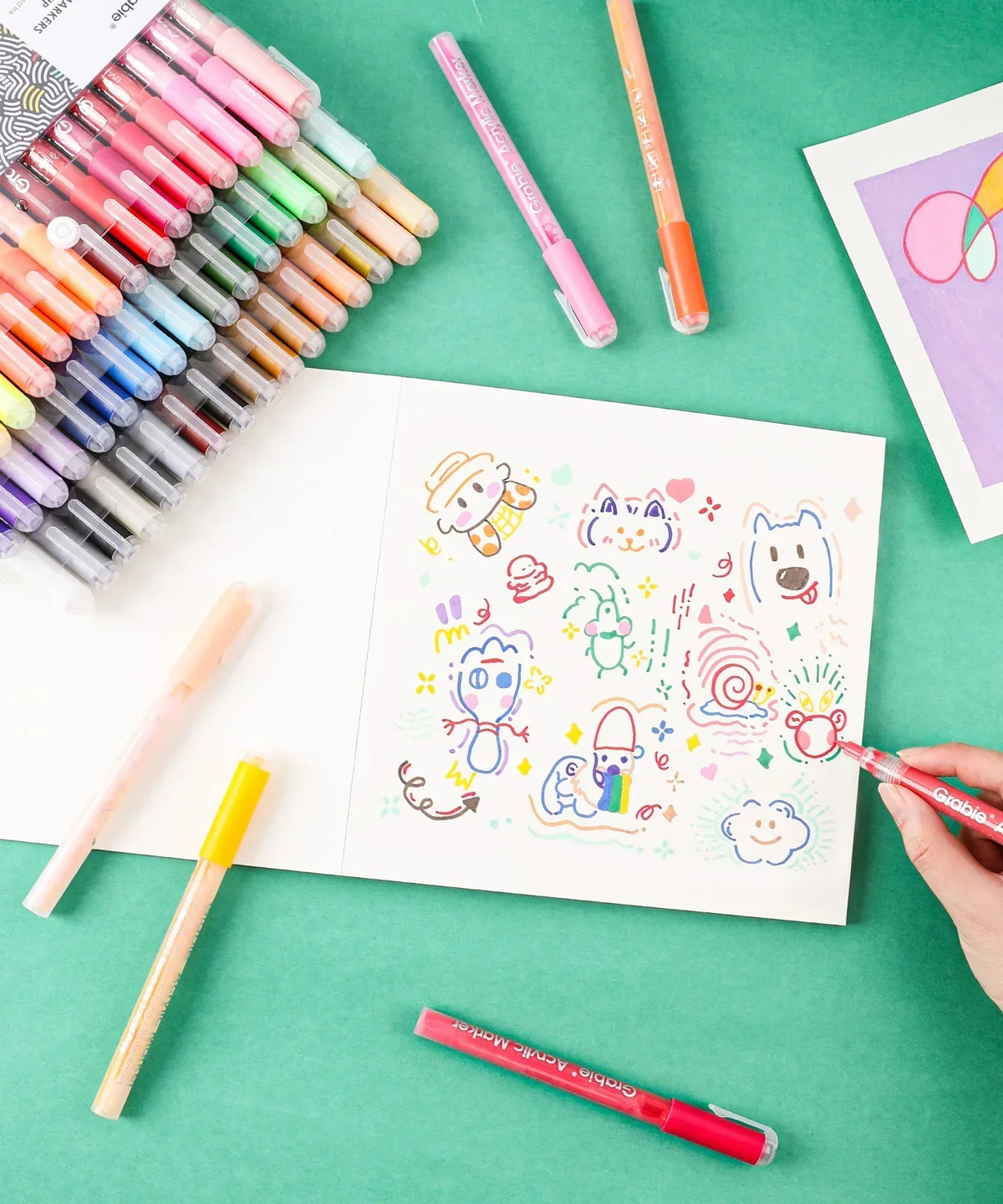 Colorful hand-drawn doodles being created with Grabie Acrylic Paint Marker Set on white paper against a green background