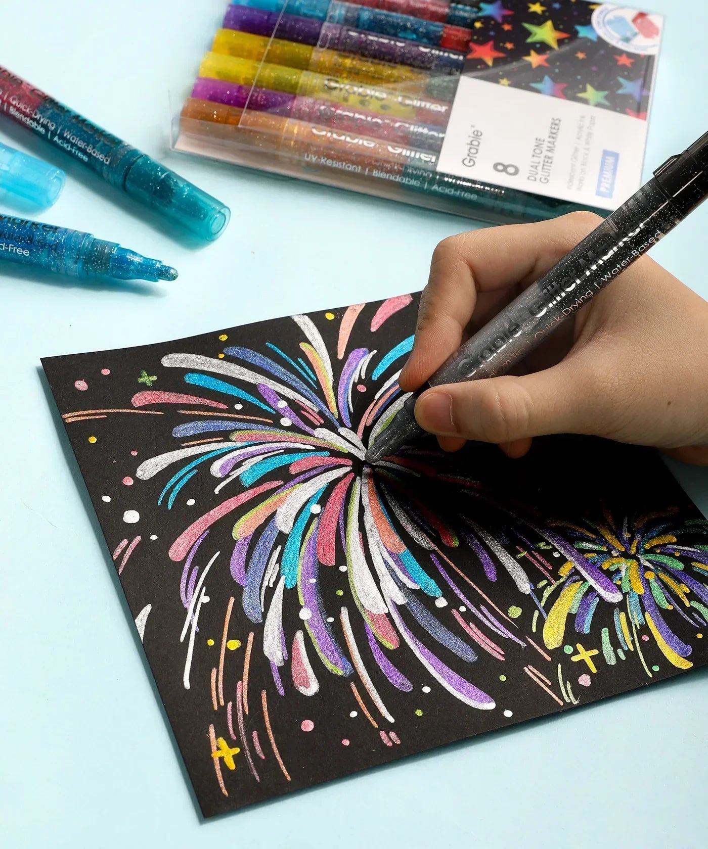 Hand using Dual Tone Iridescent Glitter Acrylic Paint Marker Set Of 8 to create colorful fireworks on black paper with pack of markers visible