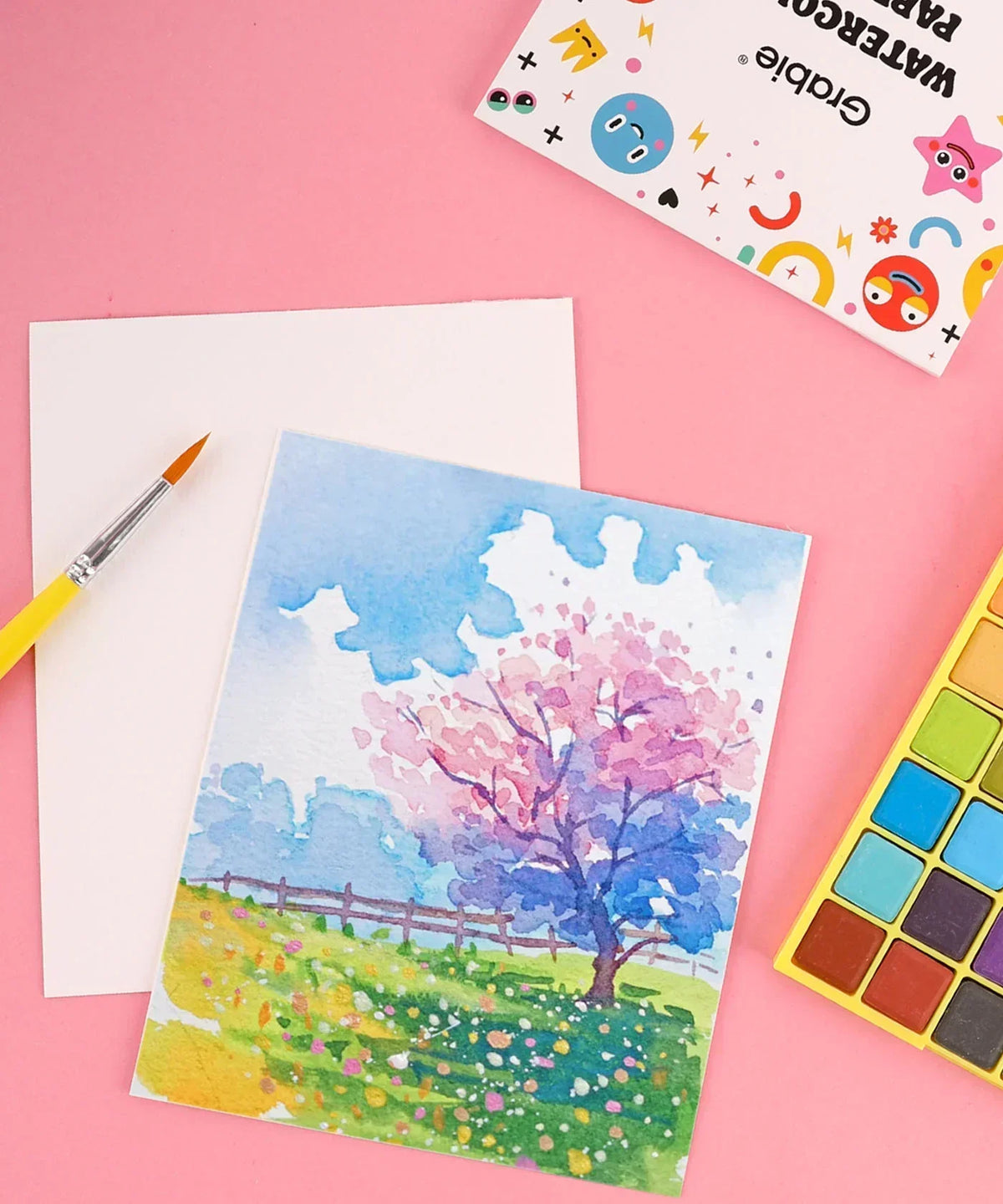 The Watercolor Beginner‘s Bundle with paintbrush, watercolor palette, and artwork of a colorful tree and landscape on pink background