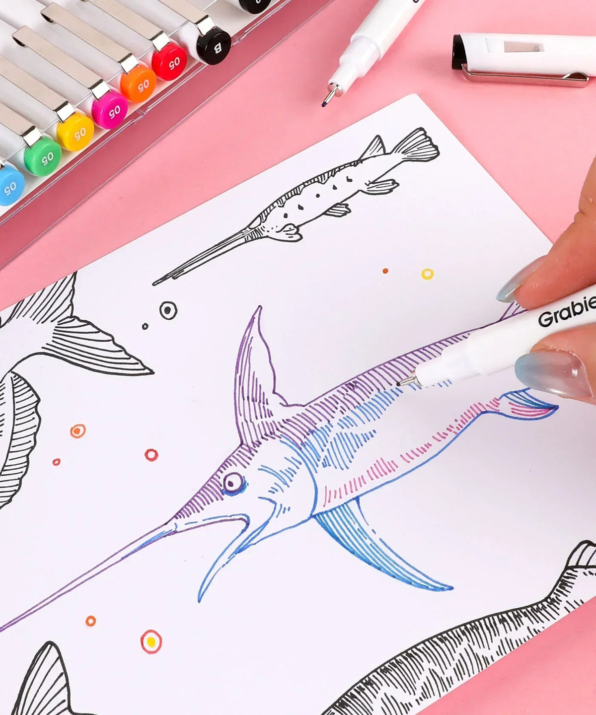 Hand using Waterproof Multiliner Drawing Pen Set Of 10 to color detailed fish illustration on white paper with art supplies nearby