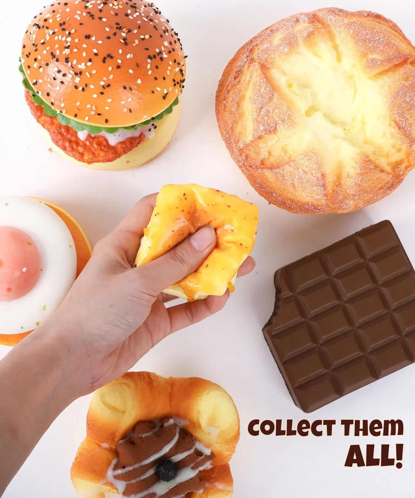 Hand squeezing yellow Grabie Ultimate Stress Relief Squishy surrounded by food-shaped squishies on a white background