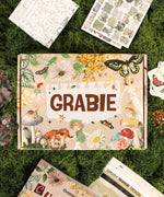 The Botanical Muse-Themed Grabie Scrapbook Club Box with floral, butterfly, and vintage art supplies on green moss background