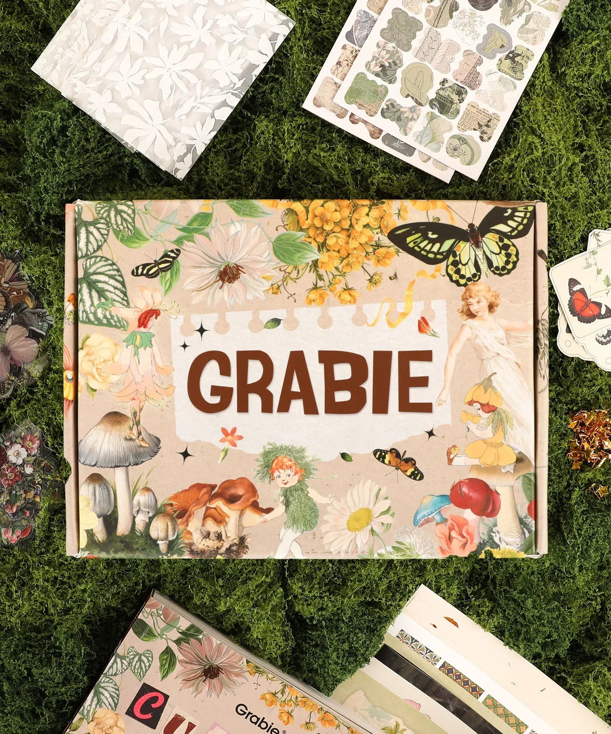 The Botanical Muse-Themed Grabie Scrapbook Club Box with floral, butterfly, and vintage art supplies on green moss background
